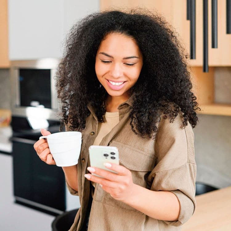 Woman in a brown-beige jacket typing on her phone and drinking coffee.