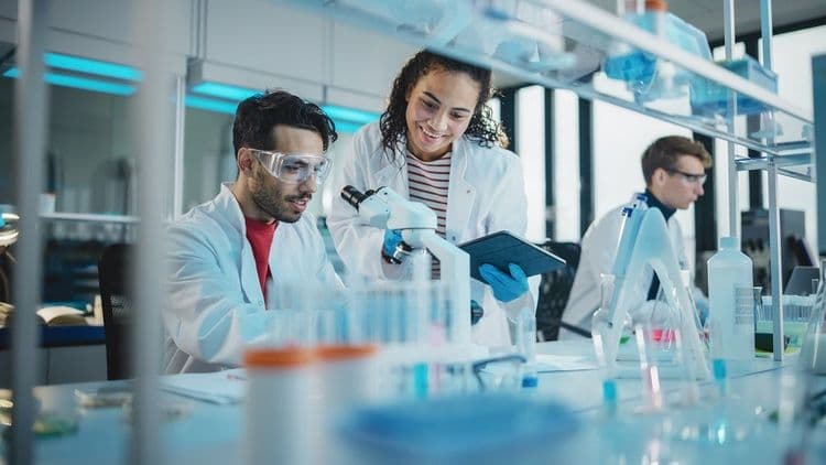 A man and woman working in a science lab looking through a microscope and taking notes.